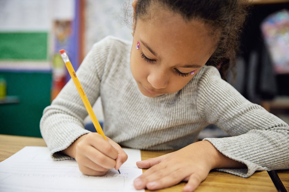 A young student sits at a desk and uses a pencil to write on white paper.