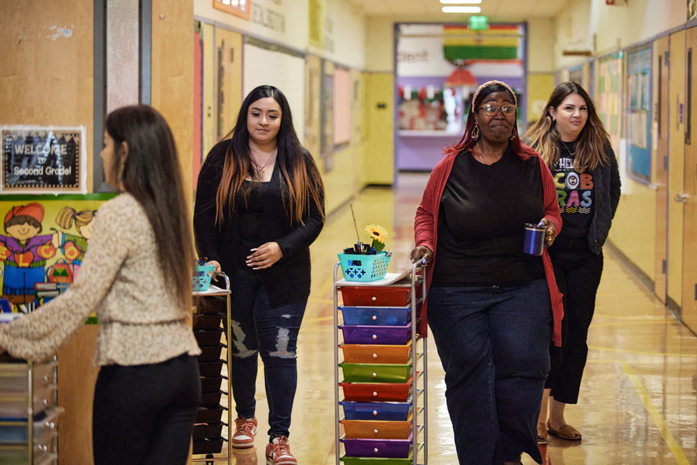 Three adults push colorful drawer carts through an elementary school hallway. A fourth adult walks behind them.