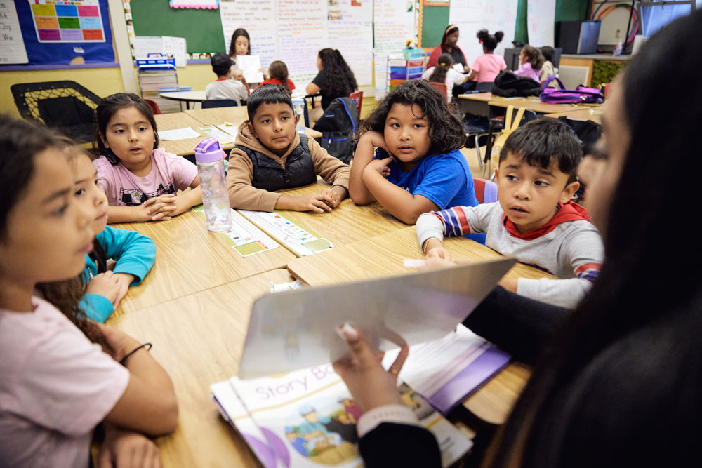 Six seated children listen to an adult teacher, who shows them a small whiteboard. In the background, two additional tables can be seen, where other adults and children are engaged in activities.