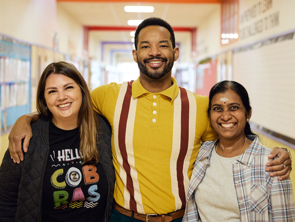 Three people stand close together, smiling. The middle person has their arms around the shoulders of the two people beside them, showing a sense of connection and camaraderie.