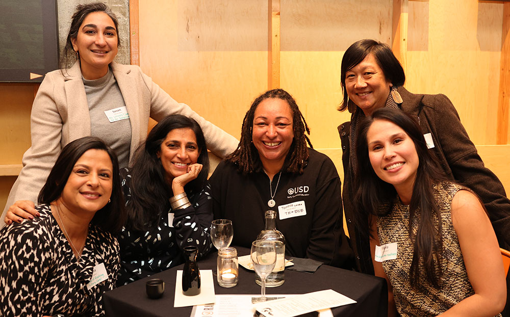 Six people smile at the camera. They are huddled around a table with wine and water glasses.