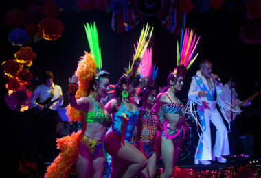 Four dancers are arm in arm wearing colorful fluorescent feathered headwear and one and two piece leotards. In the background a person with blue and white flared jumpsuit plays the guitar.