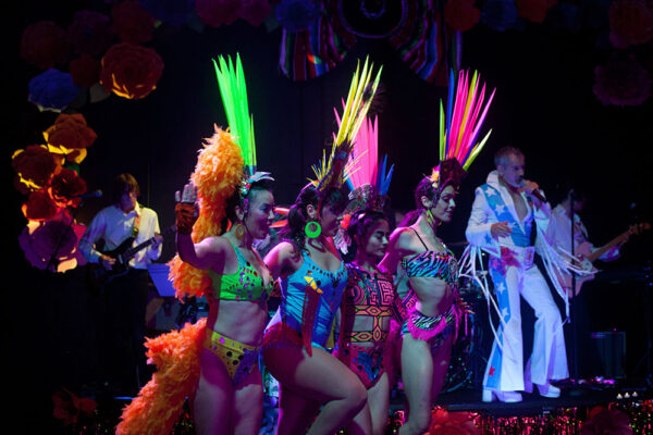 Four dancers are arm in arm wearing colorful fluorescent feathered headwear and one and two piece leotards. In the background a person with blue and white flared jumpsuit plays the guitar.