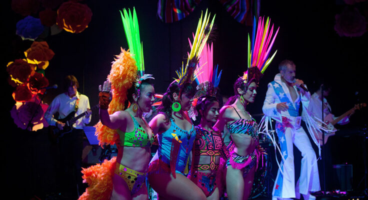 Four dancers are arm in arm wearing colorful fluorescent feathered headwear and one and two piece leotards. In the background a person with blue and white flared jumpsuit plays the guitar.