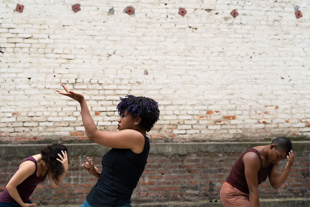 A person with short black braids with purple highlights stands in profile with their palm raised up. In the background two dancers lean toward the ground with their palms on their head.