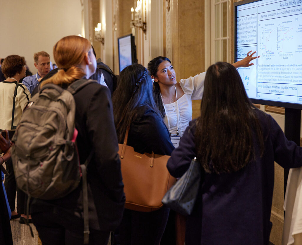 A group of people standing around a digital poster displayed on a large TV screen as the researcher points out their findings.