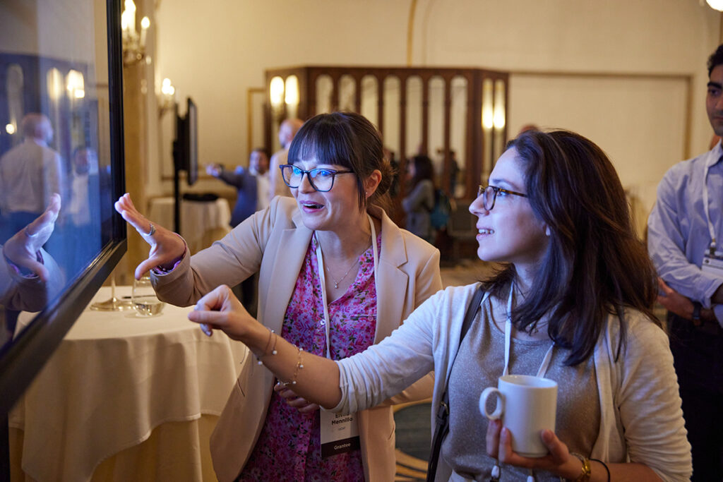 Two people animatedly discussing research while pointing at a digital poster displayed on a big TV screen at the Symposium.