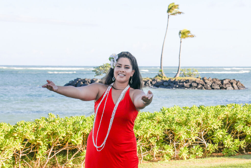 A Hula dancer wearing a bright red dress with her arms open wide and the ocean and palm trees in the background.