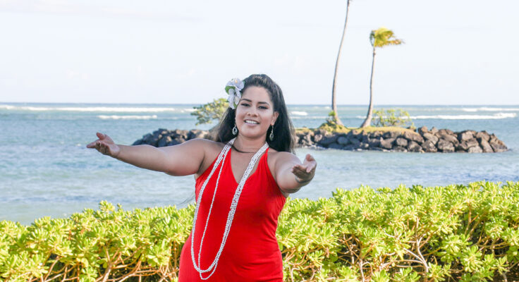 A Hula dancer wearing a bright red dress with her arms open wide and the ocean and palm trees in the background.
