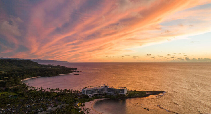 An aerial shot of the Ritz-Carlton Oʻahu, Turtle Bay, at sunset. The hotel is right on the water and the light blue sky in the background is painted with streaks of pink and orange.