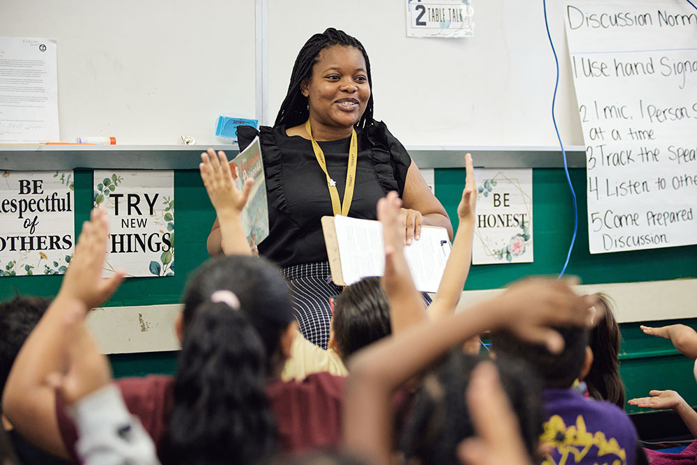 A teacher with a wide grin faces the camera and holds a book up. In front of her, a group of students are seated on the floor and eaglerly raising their hands.