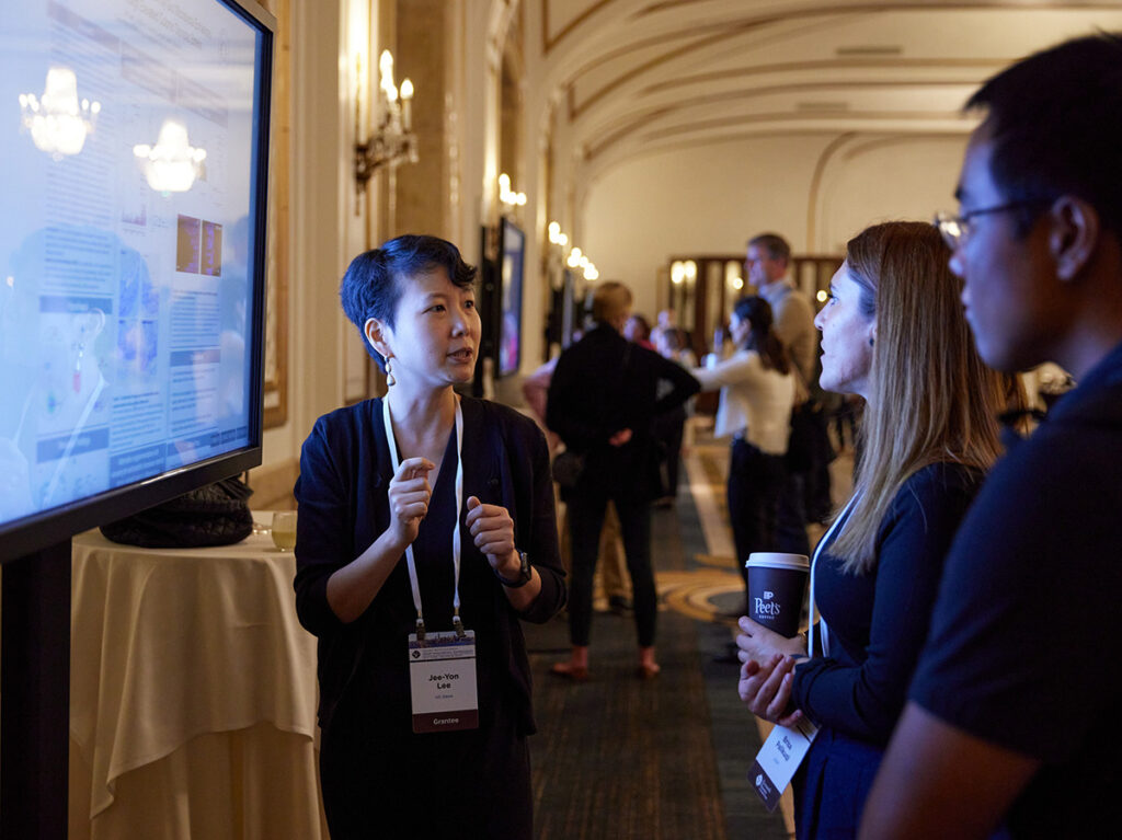 Someone animatedly presenting their research in front of a digital poster displayed on a TV screen.