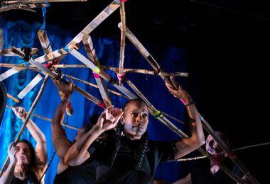 Dancers dressed in black holding up a contraption of wooden pieces held together by colorful tape.