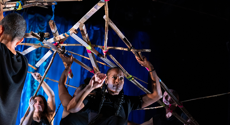 Dancers dressed in black holding up a contraption of wooden pieces held together by colorful tape.