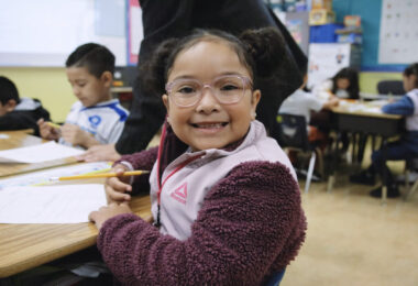 kindergarten student sits a table holding a pencil while smiling big directly at the camer