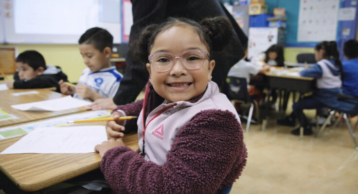 kindergarten student sits a table holding a pencil while smiling big directly at the camer