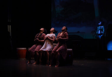 Three people sit on a bench on a darkly lit stage. Their heads tilt back and they are looking up at something not in the photo. Their hands are cupped over their chests.