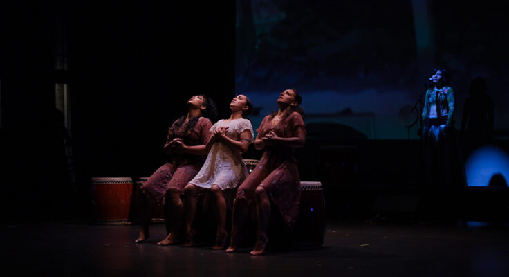 Three people sit on a bench on a darkly lit stage. Their heads tilt back and they are looking up at something not in the photo. Their hands are cupped over their chests.