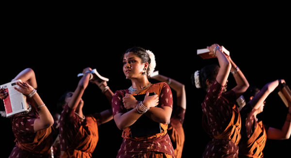 In the foreground, a hopeful dancer gazes outward, clutching a book close to her body. In the background, five other dancers are captured in motion, symbolizing struggle and labor.