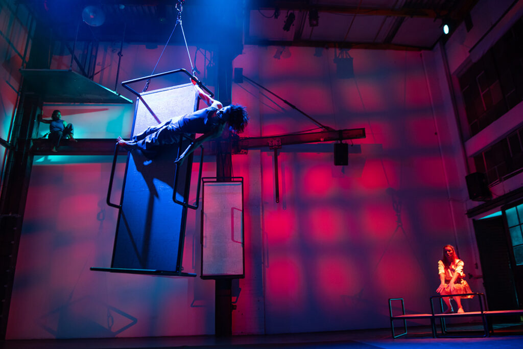 An aerial dancer in the foreground hangs from a contraption held up by a large chain.