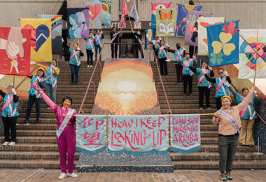 People shown on two sides of a staircase, raising multi-colored flags and smiling at the camera.