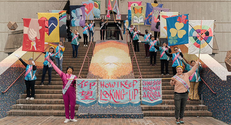 People shown on two sides of a staircase, raising multi-colored flags and smiling at the camera.