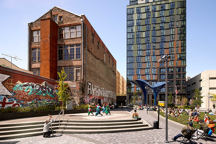 A dance performance on an outdoor stage in front of a brick building with graffiti.