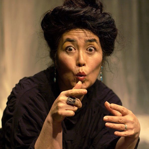 Brenda Wong Aoki, an Asian American woman, looks at the camera with an exaggerated expression on her face. She is wearing a black top and long turquoise earrings, and her dark hair is twisted into a knot on top of her head. One of her hands points toward the viewer.