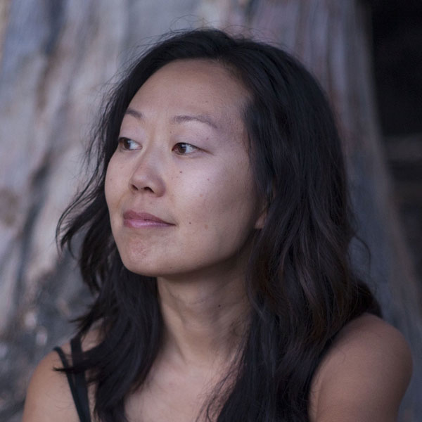 Christy, a Chinese-American woman with wavy, shoulder-length black hair looks towards left of camera. She is in front of a blurred outdoor landscape.