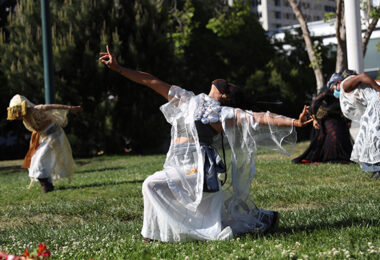 Three dancers dressed in white garments performing on a grassy field.