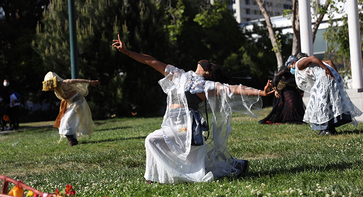 Three dancers dressed in white garments performing on a grassy field.
