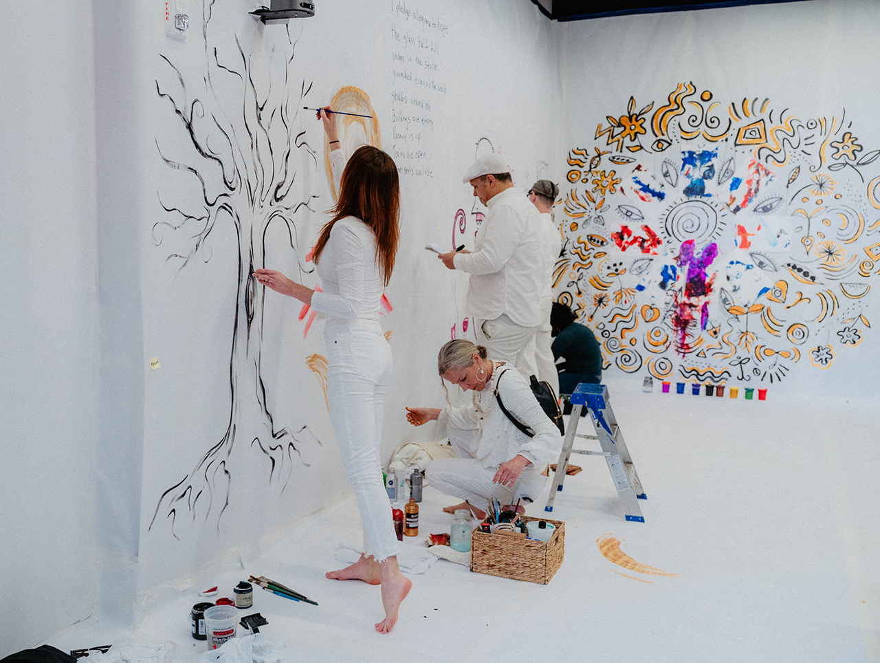 People dressed in white, adding colorful paintings to white walls in a building.