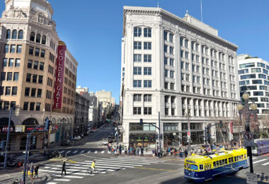 A street scene, photographed from up high, shows three prominent buildings, people walking and a blue and yellow street car.