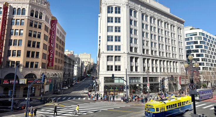 A street scene, photographed from up high, shows three prominent buildings, people walking and a blue and yellow street car.