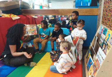 An adult sits cross legged on a rainbow striped mat and reads a book, while six children listen intently.