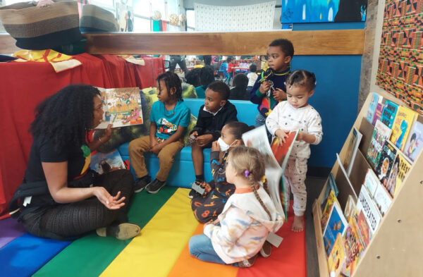 An adult sits cross legged on a rainbow striped mat and reads a book, while six children listen intently.