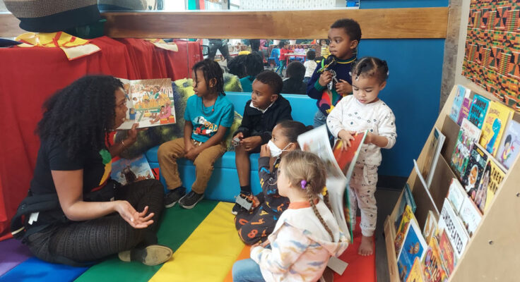 An adult sits cross legged on a rainbow striped mat and reads a book, while six children listen intently.