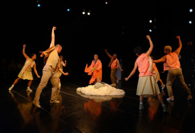 Group of dancers form a circle on a dark stage. Their right arms extend up as they look down at a pile of white spooled fabric