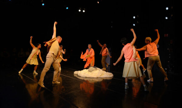 Group of dancers form a circle on a dark stage. Their right arms extend up as they look down at a pile of white spooled fabric