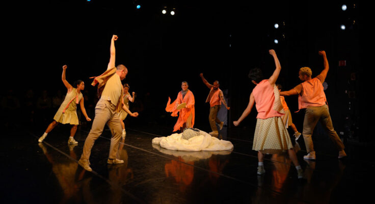 Group of dancers form a circle on a dark stage. Their right arms extend up as they look down at a pile of white spooled fabric