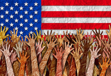 Many hands with different skin tones raised in front of the American flag.