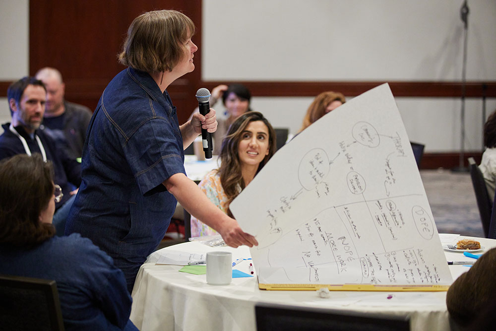 A person standing a table speaks into a microphone while flipping a large piece of paper; people at nearby tables listen attentively.