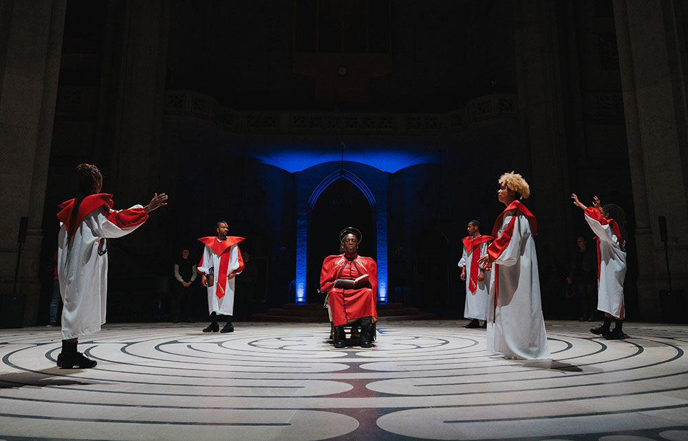 A person dressed in red sits holding a book in front of a glowing blue arch. Surrounding them are five figures in white robes with large red collars and scarves, all standing on a circular labyrinth pattern.
