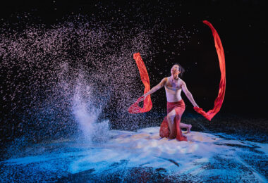 Against a dark backdrop a dancer crouches amid piles of white rice, waving long red fabric banners in each hand. In front of them, sheets of rice pour from above.