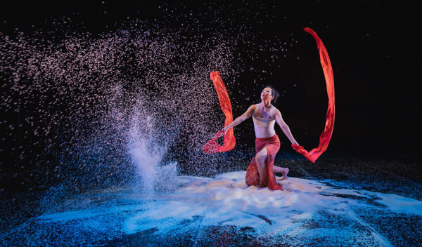 Against a dark backdrop a dancer crouches amid piles of white rice, waving long red fabric banners in each hand. In front of them, sheets of rice pour from above.