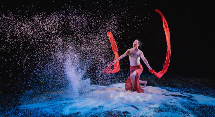 Against a dark backdrop a dancer crouches amid piles of white rice, waving long red fabric banners in each hand. In front of them, sheets of rice pour from above.