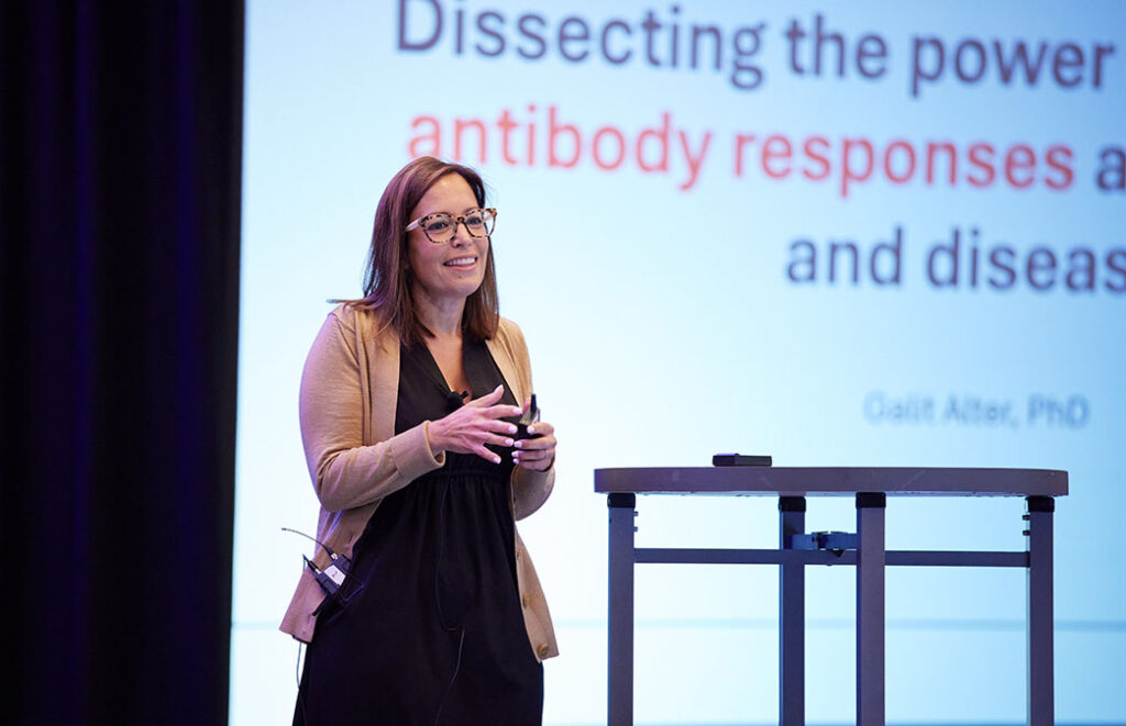 A person stands facing an audience that is out of frame. In the background half a projection is visible with the words "dissecting the power antibody responses and disease"