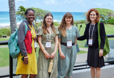 Four people stand closely posing for a photo, in the background is lush freen grass, palm trees and the ocean.