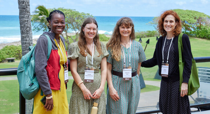 Four people stand closely posing for a photo, in the background is lush freen grass, palm trees and the ocean.