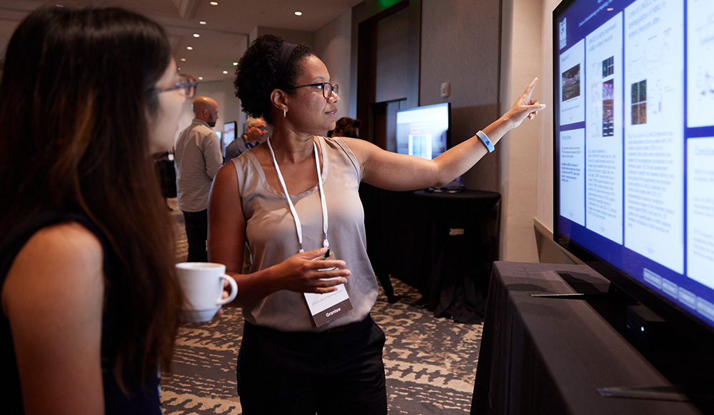 A person gestures at a digital screen while a second person listens closely.